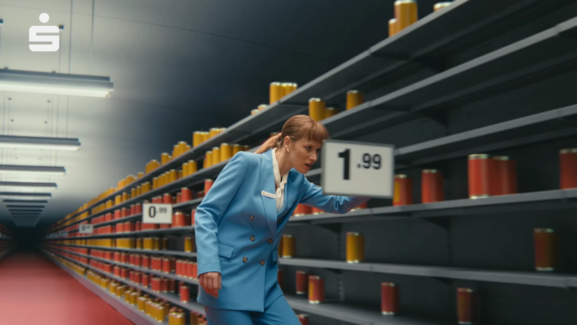 a woman walking between two shelves in a super markes and trying to pick up a can out of one shelf. in both shelves there are red and orange cans and price tags