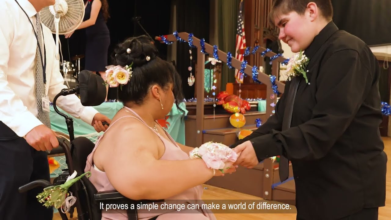 A teenage girl in a wheel chair. She is wearing a formal pink gown and corsage. A young teenage boy holds her hand.