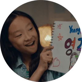 A young Asian girl smiles as she holds up a sign with the American and Korean spellings of her name.