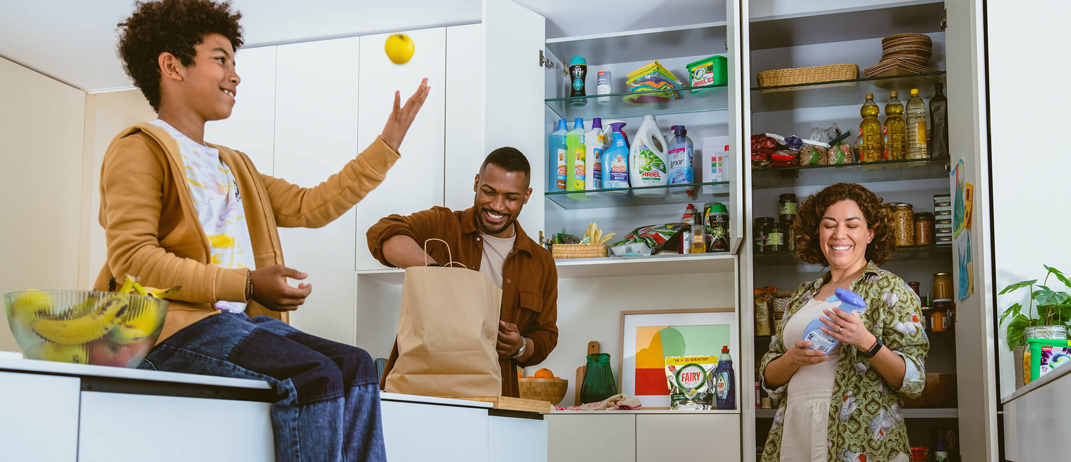 A mother and father smile as they unpack several household products into their pantry. Their son excitedly watches from the counter he is sitting on.