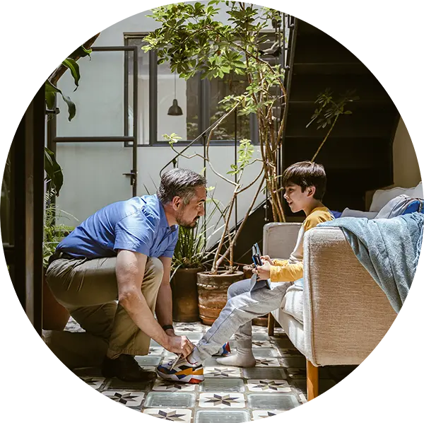 A father kneels as he ties his son’s shoe. They are in the interior courtyard of a home and surrounded by plants.
