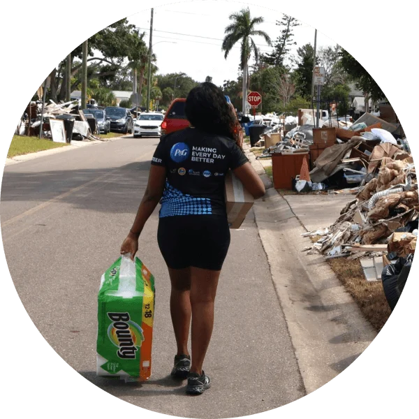 A woman in a P&G t-shirt walks on a debris-strewn street after a hurricane, carrying Bounty paper towels and a P&G disaster relief kit.