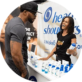 A young Black woman stands in front of a table lined with blue and white hair products. She holds one of the bottles as she engages with a young black man.
