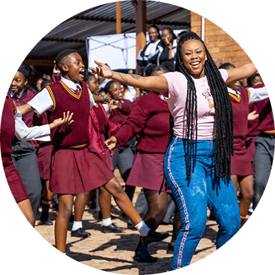 A Black woman with long, dark braided hair. She leads a larger group of female students in dance. The girls wear red and white uniforms.
