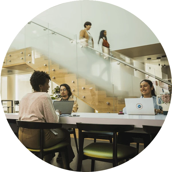 Several people sit together at various office work stations. Two women walk down a staircase in the background.