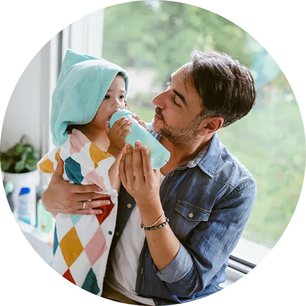 A man holds a baby wrapped in colorful towels. He holds up a bottle to the baby’s mouth.