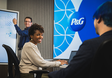 A male and female employee smile at and talk with one another while sitting at their desks at work, while a third employee writes on a white board in the background.