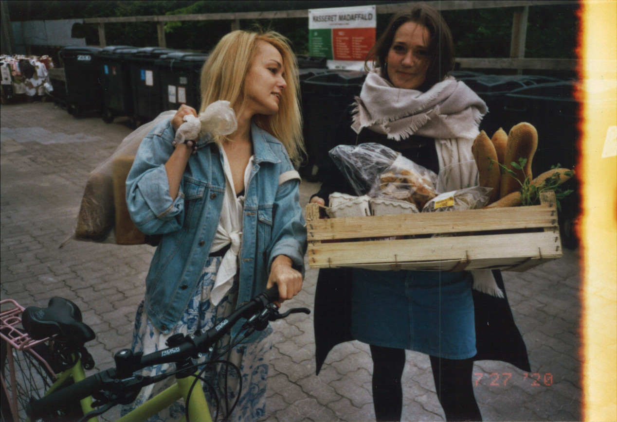 To frivillige i Food Reformers holder en pose med brød og en kurv med mad foran nogle skraldespande