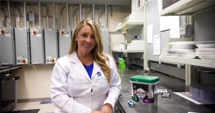 A woman with long blonde hair is wearing a white lab coat. She is standing in an industrial kitchen and smiles as she poses next to a green tub of Cascade dishwasher detergent pods that sits on the counter.