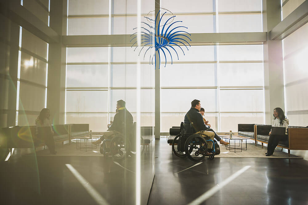 Several people sit together in a large, bright office. A man is sitting in a wheelchair, while a woman across from him sits on a bench with her laptop in her lap.