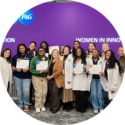 A group of women smile and pose, with some holding white certificates in their hands. Some of them wear white lab coats. Behind them, a purple screen says, “Women In Innovation” and features a round, blue P&G logo in the upper right corner.