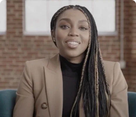 Renee Montgomery with long braided hair wearing a black top and a beige blazer smiling and looking towards the camera.