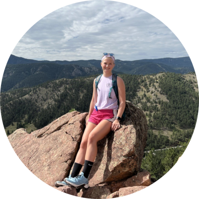 A young white woman sitting atop a large rock with mountains in the background.