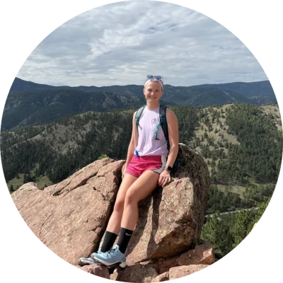 A young white woman sitting atop a large rock with mountains in the background.