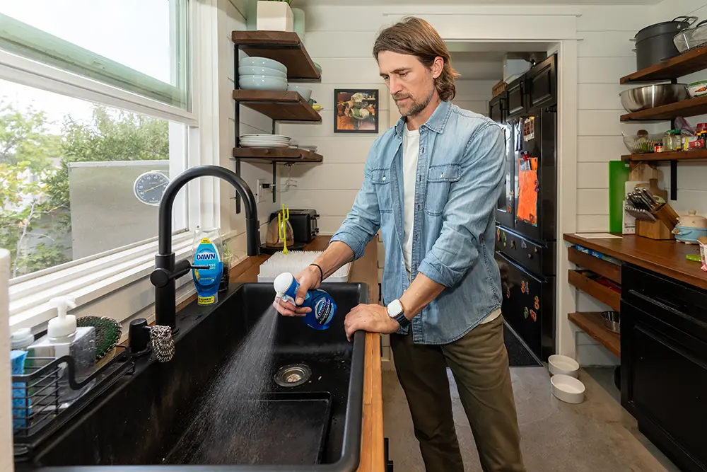A man in a denim shirt stands in a modern kitchen, spraying Dawn Powerwash into a black sink.