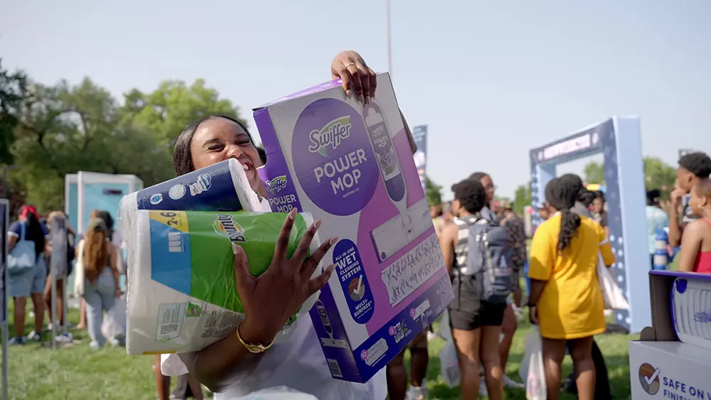A young Black woman smiles while holding packages of paper towels, toilet paper, and a Swiffer Power Mop. She is standing outside on a college campus. 