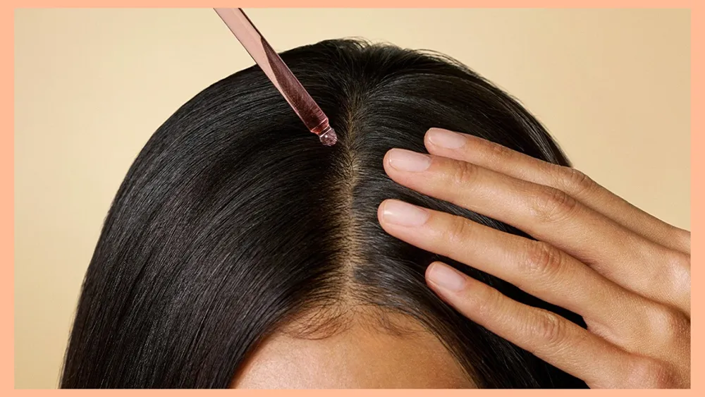 A close-up shows a dropper applying liquid to a person's dark hair part, with a hand touching their head