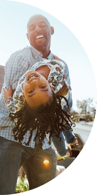 A father stands in his front yard and laughs while holding his smiling daughter who is leaning backwards in his arms