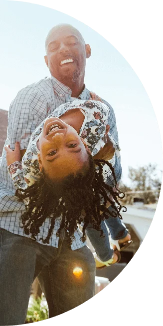 A father stands in his front yard and laughs while holding his smiling daughter who is leaning backwards in his arms