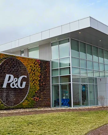 Photograph of a P&G facility featuring large glass windows and a vertical garden wall with a prominent white "P&G" logo. The setting includes a grassy foreground and cloudy sky.