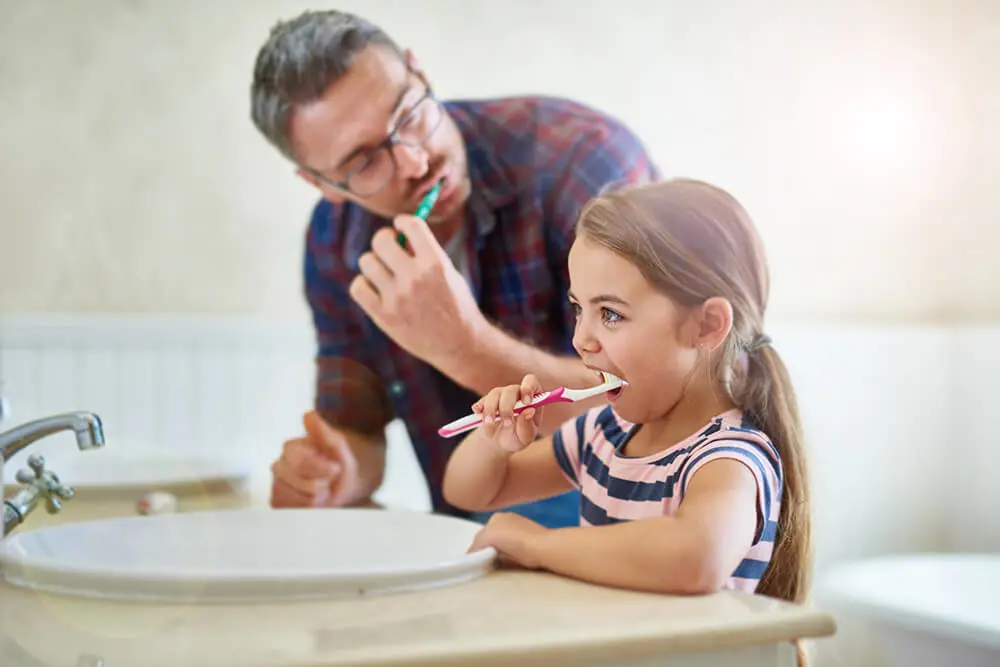 Father coaching daughter on healthy brushing habits.