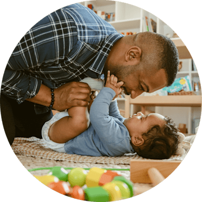 A man in a blue shirt joyfully smiles at a baby who is wearing a diaper and playfully grabbing the man’s face.