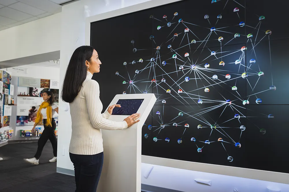 A women with long dark hair is dressed in a long, white knit sweater and black pants. She stands in front of an interactive kiosk that projects an interconnected web of P&G brand logos.