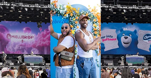 Two men are standing back to back, smiling and facing the camera, with a heart-shaped flower arrangement behind them.