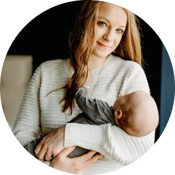 A woman with long hair is wearing a white sweater and is holding a baby in her arms. She is smiling towards the camera.