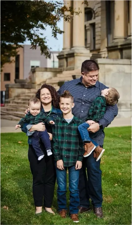 Woman with dark curly hair, stands holding her youngest son, while her husband on the right holds another son who is looking up at him. Their oldest boy stands between them. All are wearing blue plaid shirts and smiling.