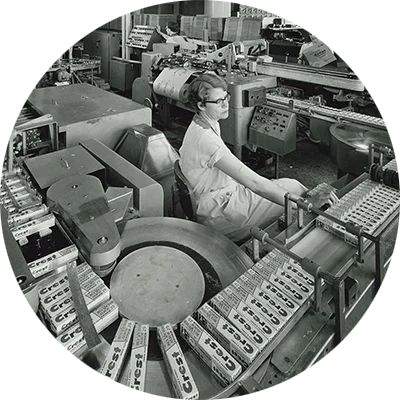 Archival image of a factory where women are packaging Crest toothpaste. Conveyors transport the boxes