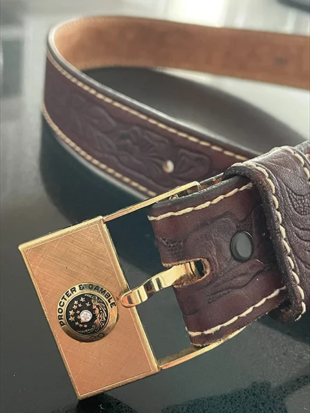 A brown belt with a gold buckle and P&G logo emblem.