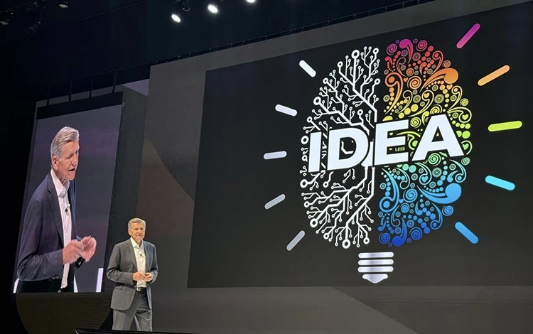 A white man in a grey business suit and white shirt stands on a stage. An illustrated of a brain, in the shape of a light bulb, and the white text that says "idea" are projected onto a large screen in the background.