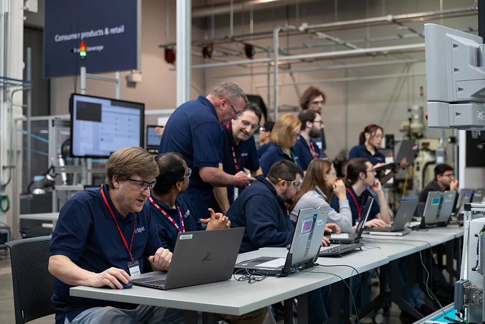 Participants at a table, each with a laptop, and several others standing behind them