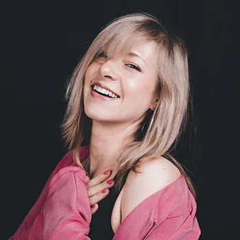 Headshot of a woman with light skin, blond hair, a big smile and pink shirt. She has her hands over her heart, and the background is black.