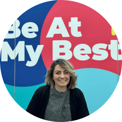 A woman with short blonde hair smiles. She stands in front of a colorful backdrop with the words “be at my best” written in white text.