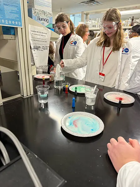 Two young women wear lab goggles and white lab coats. They stand over a black table and hold a white cloth over a beaker filled with liquid. Paper plates holding colored liquids sit on the counter.