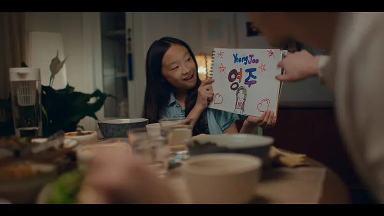 A young Asian girl with long hair and a blue shirt sits at the dinner table with her family holding a drawing of her name “Yeong Joo.”