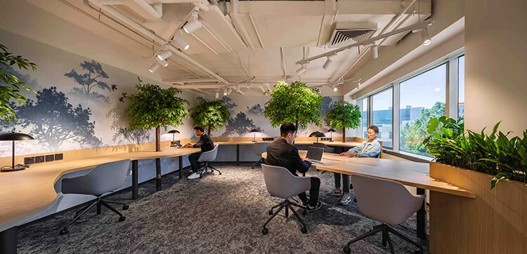 Room featuring natural brown color tones and warm light throughout. A curved wood desk lines the walls, while green plants and trees are placed throughout the room, with a large window letting in natural light.