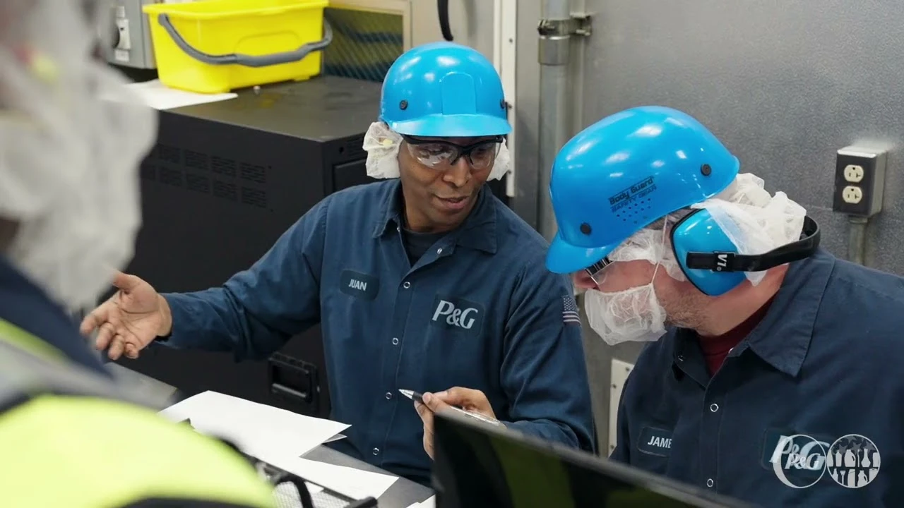 Two factory workers wearing blue hard hats, safety glasses, and protective gear discussing work inside an industrial facility