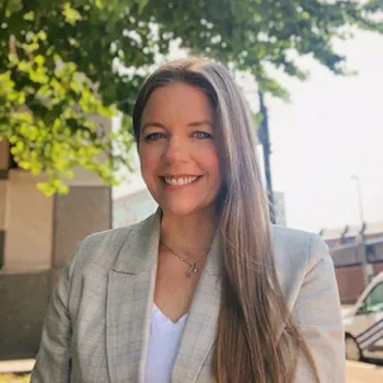 Headshot of a smiling woman standing outside under a tree. She has light skin and very long light brown hair and is wearing a tan jacket and white shirt. 