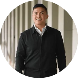 A man with dark hair, in a black jacket, smiles as he stands in the hallway of an office.