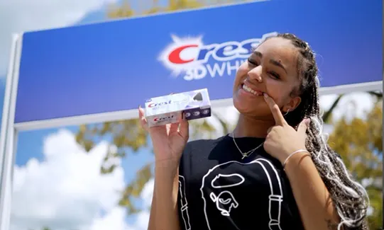 A female student points at her white teeth and smiles while holding a Crest product box, standing in front of a Crest 3D White banner backdrop. 