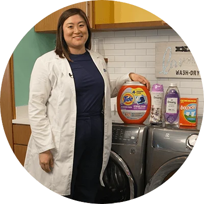 An Asian woman with shoulder length wears a white lab coat. She stands next to a washer and dryer, with four bottles of various laundry products lined across the top.