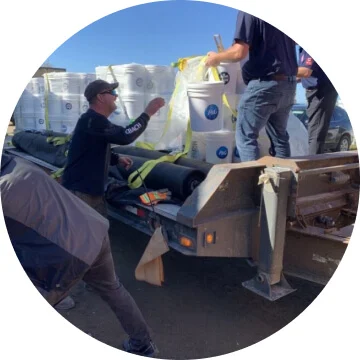 White buckets with the P&G logo on it are being unloaded from a truck bed by two people.