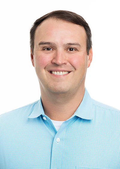 A white man with short brown hair in a blue button up shirt smiles directly at the camera.