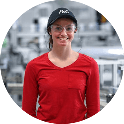 A woman in a long sleeved red shirt, black baseball cap, safety goggles and ear plugs. A manufacturing plant blurred in the background.