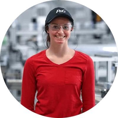 A woman in a long sleeved red shirt, black baseball cap, safety goggles and ear plugs. A manufacturing plant blurred in the background.