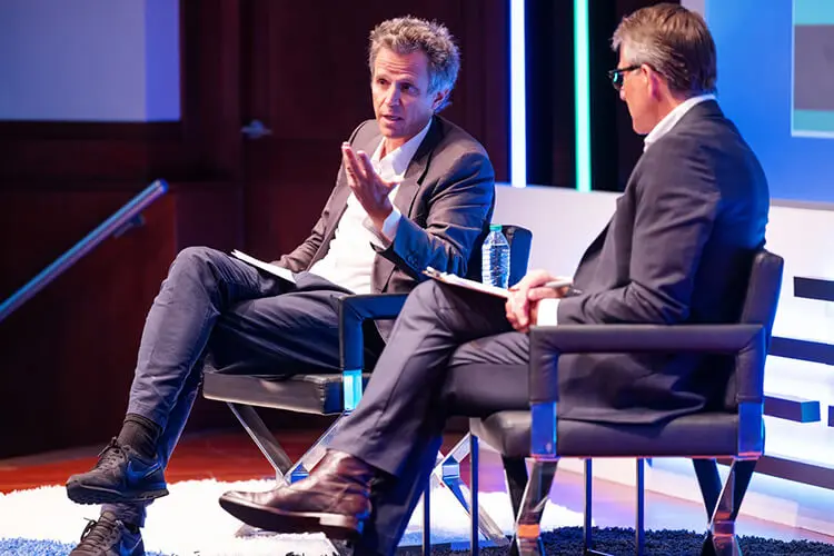 Two mean wearing business suits sit on a stage for a conversation.