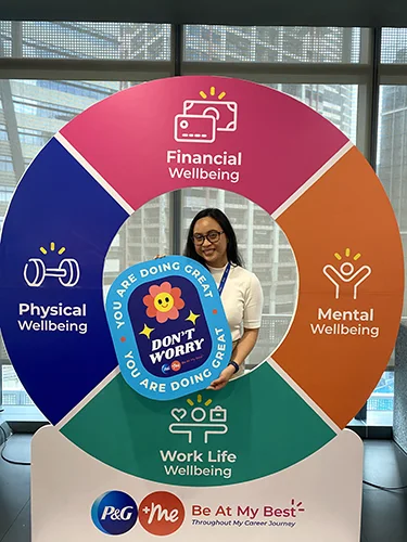 A smiling woman peeks through a large, colorful sign promoting "Wellbeing." It lists Mental, Financial, Work Life, and Physical Wellbeing. She holds a "Don't Worry" sign.